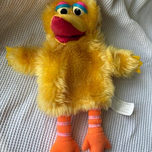 Puede incluir: Una marioneta de mano de peluche amarilla con forma de pájaro, con pelaje largo y desgreñado, un pico rojo y párpados azules y rosas. La marioneta tiene dos patas naranjas con rayas horizontales rosas y pies de tres dedos, con un mechón.