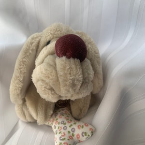 以下が含まれることがあります： 茶色の鼻と花柄の骨を持つ、ベージュのぬいぐるみ犬のおもちゃ。おもちゃには「25719」という番号のタグが付いています。