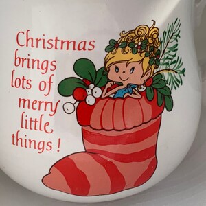 May include: White ceramic mug with a red and white striped stocking and a cartoon fairy with blonde hair and a blue dress. The text on the mug reads "Christmas brings lots of merry little things!"