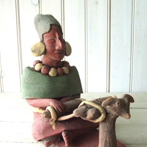 May include: A terracotta figurine of a woman wearing a green tunic and a beaded necklace, holding a small animal in her lap. The figurine is painted in shades of brown, green, and yellow.