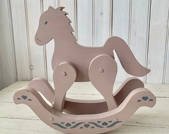 May include: A wooden rocking horse painted in a light pink color with a gray painted design on the base. The horse has a simple design with a mane and tail.