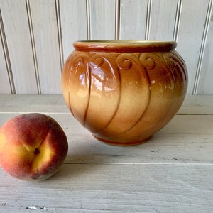 May include: A brown glazed ceramic planter with a decorative design. The planter has a rounded shape and a wide opening. It is sitting on a white wooden surface next to a peach.