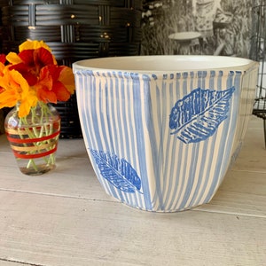 May include: A white ceramic planter with blue and white stripes and blue leaf designs. The planter is sitting on a white wooden surface.