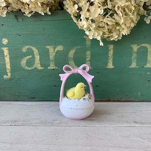 May include: A small, white and pink Easter egg-shaped container with a yellow chick inside. The container has a pink bow handle. The background includes a green wooden surface with the word "jardin" and dried flowers.