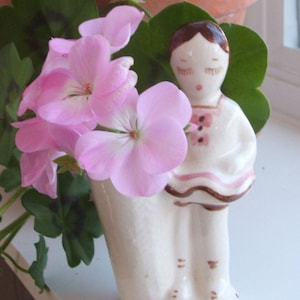 May include: A white ceramic figurine of a girl wearing a pink and brown dress. The figurine is holding a bouquet of pink geraniums.