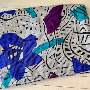 May include: A folded piece of fabric featuring a light gray base with a textured waffle-like pattern. The surface is decorated with a bold graphic print consisting of abstract shapes in vibrant royal blue, deep purple, and bright teal, accented by black lines.