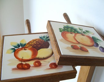 May include: Two square trivets with wooden handles and frames. Each trivet features a white tile with a painted fruit design. One shows a pineapple, apple, and cherries. The other has peaches, grapes, and plums.