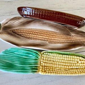 May include: Three ceramic corn-shaped dishes. One dish is dark brown with a textured kernel pattern. Another is a long, beige and brown dish shaped like a corn cob with husks. The third is a bright green and yellow dish shaped like a corn cob.