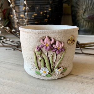 May include: A white ceramic planter with a raised design of purple and pink irises and white daisies. A yellow butterfly is on the side of the planter.