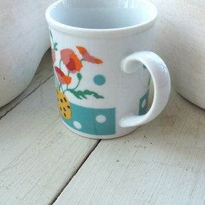Puede incluir: Taza de cer&aacute;mica blanca con un dise&ntilde;o floral azul, verde y amarillo. La taza tiene un asa blanca y est&aacute; sobre una superficie de madera blanca.