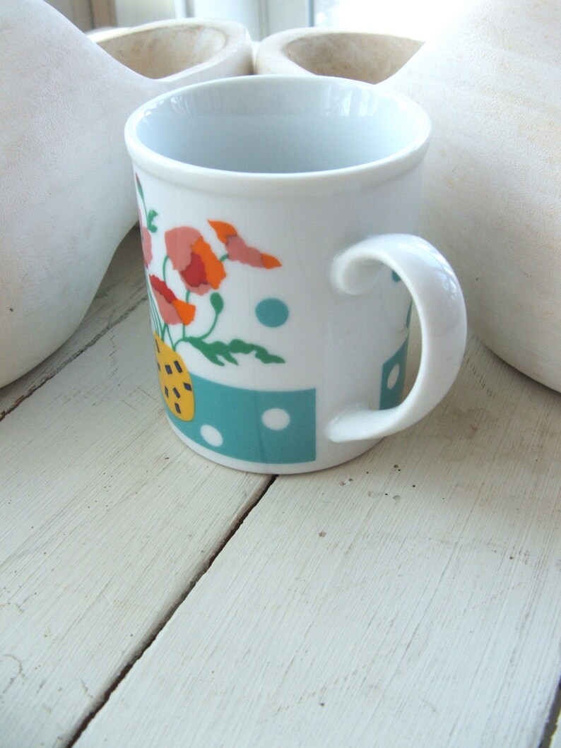 Puede incluir: Taza de cer&aacute;mica blanca con un dise&ntilde;o floral azul, verde y amarillo. La taza tiene un asa blanca y est&aacute; sobre una superficie de madera blanca.