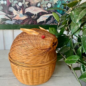 May include: A woven wicker basket shaped like a rabbit with a red button nose. The basket is made of natural brown wicker and has a round shape.