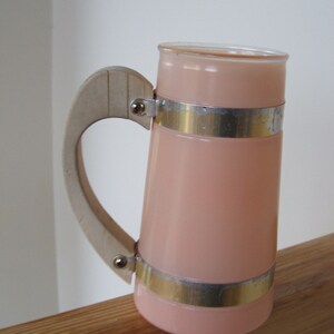 May include: A pink glass pitcher with a white handle and silver metal bands. The pitcher has a rounded shape and a wide mouth.