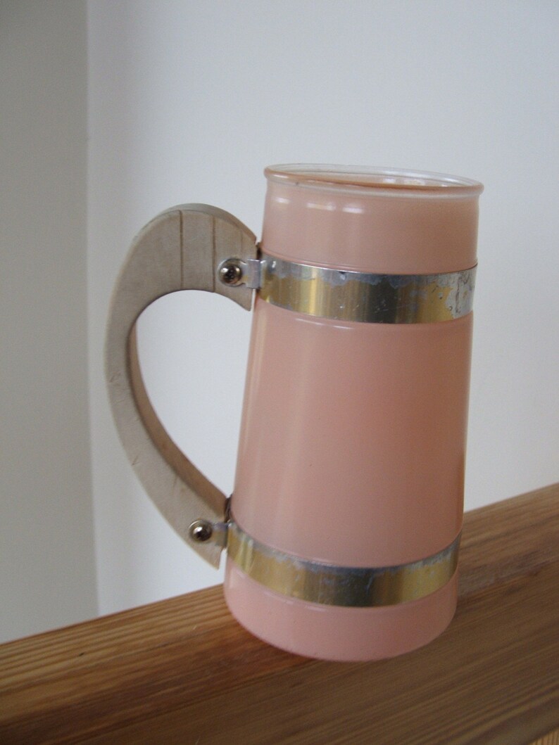 May include: A pink glass pitcher with a white handle and silver metal bands. The pitcher has a rounded shape and a wide mouth.