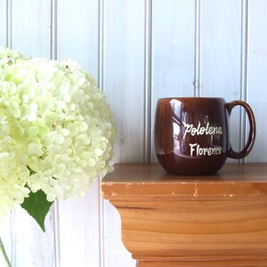 May include: A brown ceramic mug with a white inscription that reads "Pololena Florence". The mug is sitting on a wooden shelf.