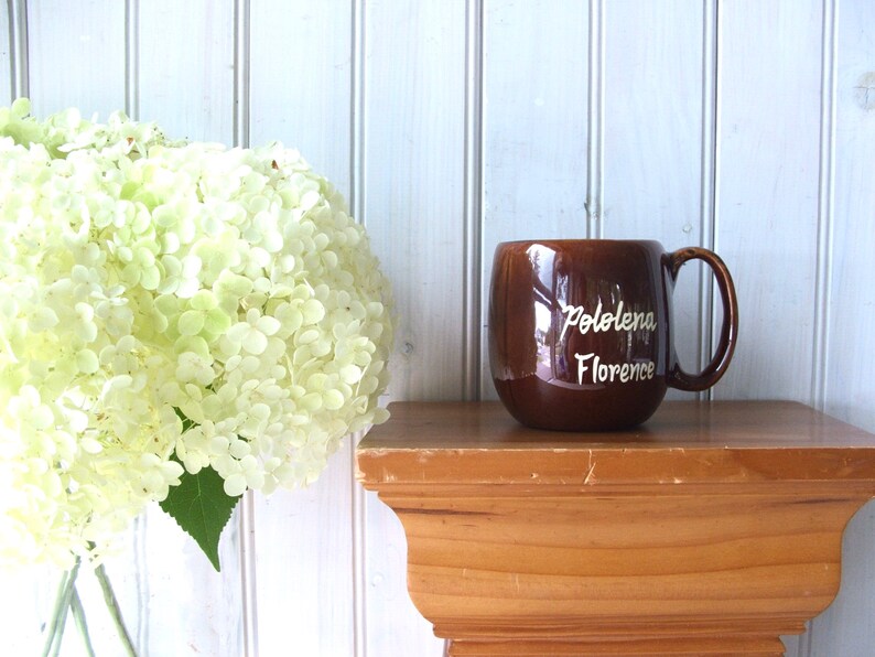 May include: A brown ceramic mug with a white inscription that reads "Pololena Florence". The mug is sitting on a wooden shelf.