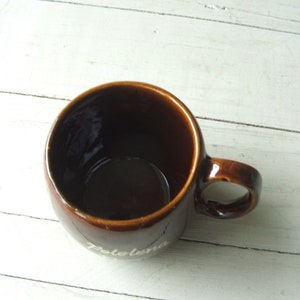 May include: A brown ceramic mug with a white inscription that reads "Zolema". The mug has a rounded shape and a handle.