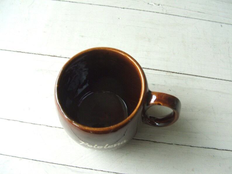 May include: A brown ceramic mug with a white inscription that reads "Zolema". The mug has a rounded shape and a handle.
