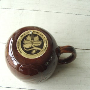May include: Brown ceramic mug with a gold logo that reads "KavaCraft Hawaii, Ltd. 894 Queen Street, Honolulu, Hawaii". The logo features a stylized flower with a stem.