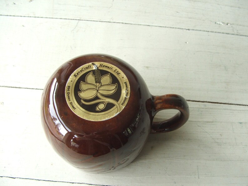 May include: Brown ceramic mug with a gold logo that reads "KavaCraft Hawaii, Ltd. 894 Queen Street, Honolulu, Hawaii". The logo features a stylized flower with a stem.