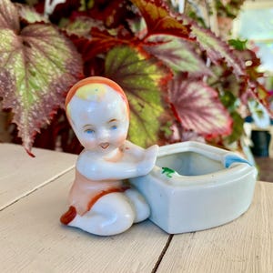 May include: A vintage porcelain figurine of a baby in a seated position, with a peach-colored outfit and a white basin. The baby has blue eyes and a red-orange cap. The basin is white with a blue accent and a green frog.