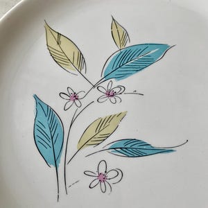 May include: White ceramic plate with a floral design. The design features stylized teal and yellow leaves with pink flowers. The plate has a simple, elegant aesthetic, suitable for serving food or as a decorative piece.