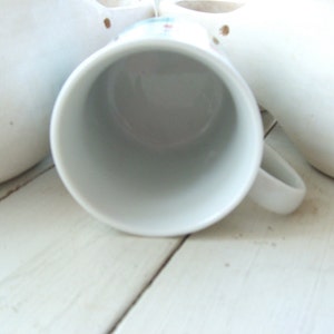 Puede incluir: Taza de caf&eacute; de cer&aacute;mica blanca con asa redondeada. La taza est&aacute; vac&iacute;a y se encuentra sobre una superficie blanca.