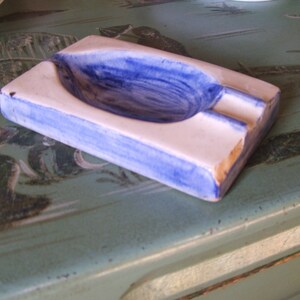 May include: A white ceramic ashtray with a blue rim and a blue interior. The ashtray is rectangular and has a rounded, oval-shaped depression in the center.