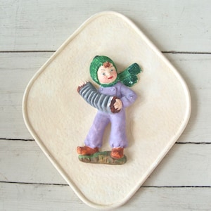 May include: A white ceramic plaque with a raised image of a child wearing a green hat and purple clothing playing an accordion. The plaque has a textured surface.