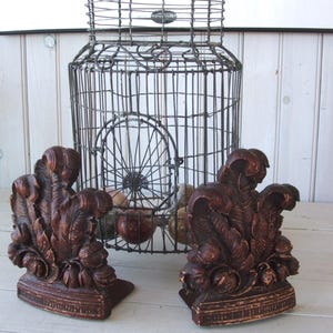 May include: Two dark brown wooden bookends with a leafy design. The bookends are sitting on a white wooden surface in front of a wire cage.
