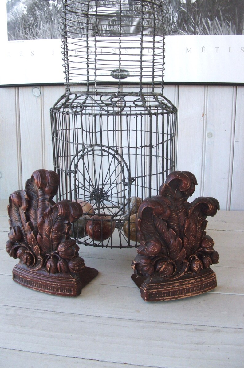 May include: Two dark brown wooden bookends with a leafy design. The bookends are sitting on a white wooden surface in front of a wire cage.