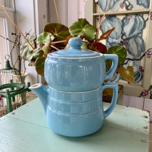 May include: A two-piece, light blue ceramic teapot set. The teapot has a lid and a spout, with a handle on each piece. The set is stacked on a weathered, light blue wooden surface, with greenery in the background.