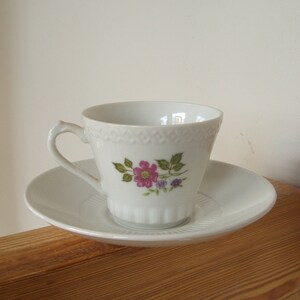 Pode incluir: Uma ch&aacute;vena de ch&aacute; e um pires de porcelana branca com um desenho floral. A ch&aacute;vena tem uma pega delicada e uma borda festonada. O pires &eacute; branco liso.