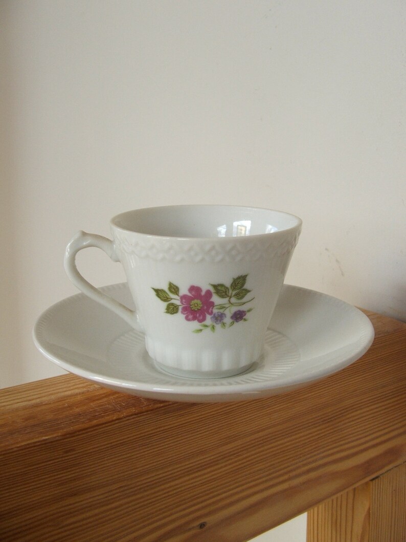 Pode incluir: Uma ch&aacute;vena de ch&aacute; e um pires de porcelana branca com um desenho floral. A ch&aacute;vena tem uma pega delicada e uma borda festonada. O pires &eacute; branco liso.