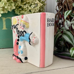 May include: A white ceramic planter shaped like a book with a pink spine. The front of the planter features a colorful illustration of a clown holding a tambourine. The text "BABY BOOK" is printed on the spine.