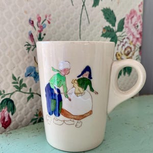 May include: A cream-colored ceramic mug featuring a hand-painted illustration of two figures wearing traditional Dutch-style clothing, including wooden clogs, a white cap, and a blue bonnet, while standing in a field with a small yellow object.