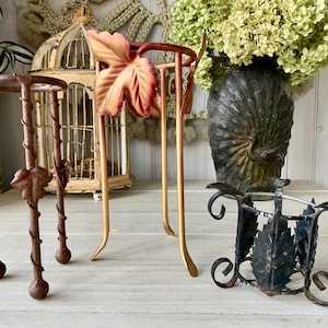 May include: An assortment of decorative metal plant stands in various styles. The stands feature leaf and vine designs, with colors including bronze, gold, and black. The stands are of varying heights and shapes, suitable for displaying small plants or decorative items.