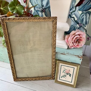 May include: Two antique gold-toned picture frames. The larger frame contains a document with faded text. A smaller frame displays a pink rose illustration. The frames are placed on a white wooden surface, with a pink rose and greenery in the background.