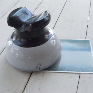 May include: A white and black ceramic insulator with the number 91 and the letter ME printed on the bottom. The insulator is sitting on a white wooden surface.