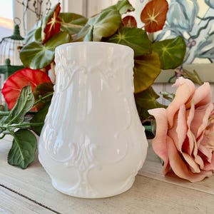 May include: A white, ornate vase with a detailed embossed design. The vase is surrounded by greenery and a pink rose, creating a vintage aesthetic. The vase's shape is slightly flared, and the embossed pattern adds a touch of elegance.