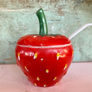May include: A ceramic strawberry-shaped container with a green stem and a white spoon. The container is red with white and yellow accents, resembling strawberry seeds. The spoon rests on the rim of the container.
