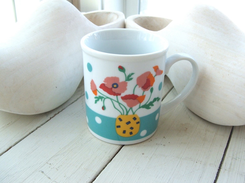 Puede incluir: Taza de cer&aacute;mica blanca con un patr&oacute;n de lunares azules y blancos y un dise&ntilde;o floral con flores rosas, rojas, amarillas y verdes. Las flores est&aacute;n en un jarr&oacute;n amarillo y negro.