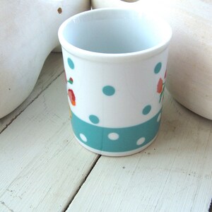 Puede incluir: Una taza de cer&aacute;mica blanca con un patr&oacute;n de lunares turquesa y blanco y un dise&ntilde;o floral. La taza est&aacute; sobre una superficie de madera blanca.