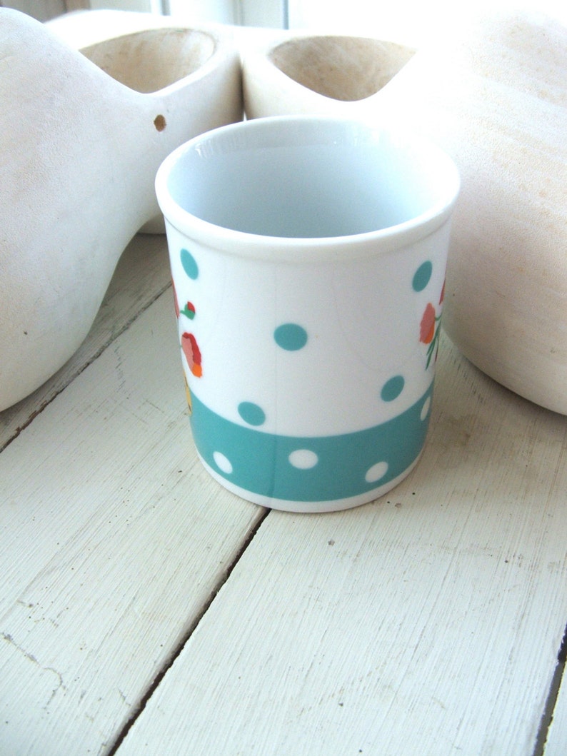 Puede incluir: Una taza de cer&aacute;mica blanca con un patr&oacute;n de lunares turquesa y blanco y un dise&ntilde;o floral. La taza est&aacute; sobre una superficie de madera blanca.