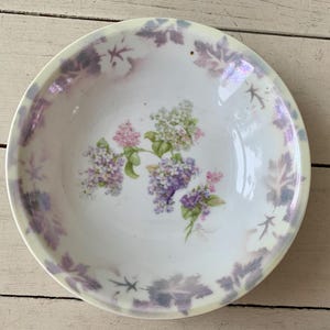 May include: A round, white ceramic bowl with a floral design. The bowl features a lilac and leaf pattern around the rim and a cluster of lilac flowers in the center. The bowl has a soft, iridescent sheen.
