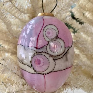 May include: Pink and white oval Christmas ornament with a gold-colored metal loop for hanging. The ornament has a decorative design with swirls and lines in black and pink. The ornament is hanging on a white Christmas tree.