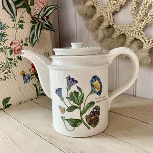 May include: White ceramic teapot with a floral and butterfly design. The teapot features a curved spout, a handle, and a lid. The floral design includes blue flowers and green leaves. The teapot is sitting on a white wooden surface.