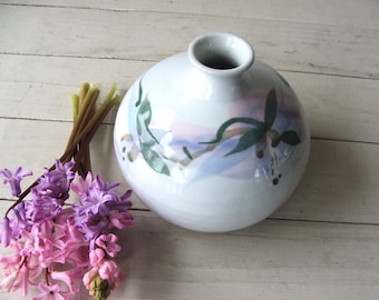 May include: White ceramic vase with a round body and a narrow neck. The vase is decorated with a hand-painted design of green leaves and purple flowers.