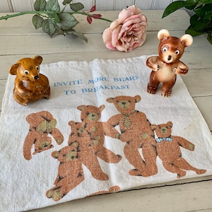 May include: A white tea towel with a brown bear design and the text "Invite more bears to breakfast". There are several brown bears printed on the towel, and two brown ceramic bear figurines are on top of the towel.