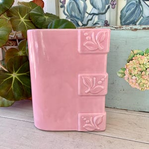 May include: A rectangular, light pink ceramic vase with three decorative leaf designs. The vase is a solid color with a glossy finish. The vase is suitable for displaying flowers or as a decorative item.